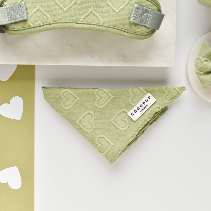 A Luxe Bandana - Sage Heart with a textured heart pattern lies flat on a white surface. Lightweight and breathable, it features a small label reading "Cocopup London." Various heart-shaped cutouts and fabric items in matching colors surround the bandana from the Cocopup range.