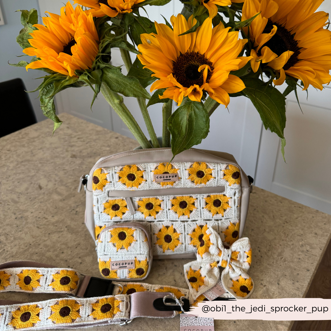 A beige handbag adorned with a sunflower pattern sits on a counter, complemented by sunflower-themed accessories and the Bag Strap - Sunflower Patch from Cocopup London. A bouquet of fresh sunflowers in the background adds a bright touch to the scene. The Instagram handle @obi1_the_jedi_sprocker_pup is visible.