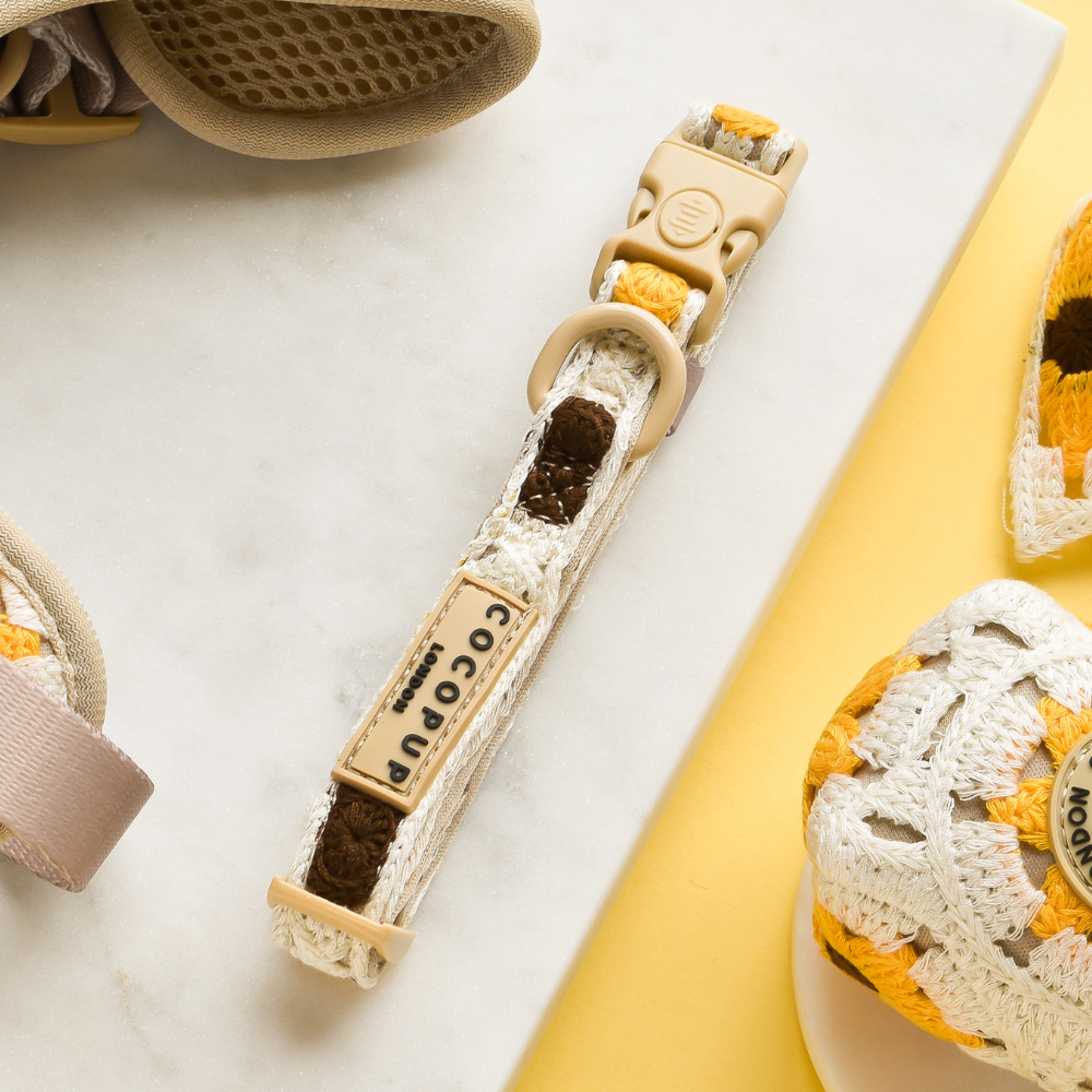 A cream and yellow woven baby pacifier clip with a beige plastic clasp lies on a white marble surface. The clip, adorned with a "Cocopup London" label, is surrounded by parts of baby shoes woven in similar colors, all set against the Sunflower Patch Adjustable Neck Harness, Lead & Collar Bundle backdrop.