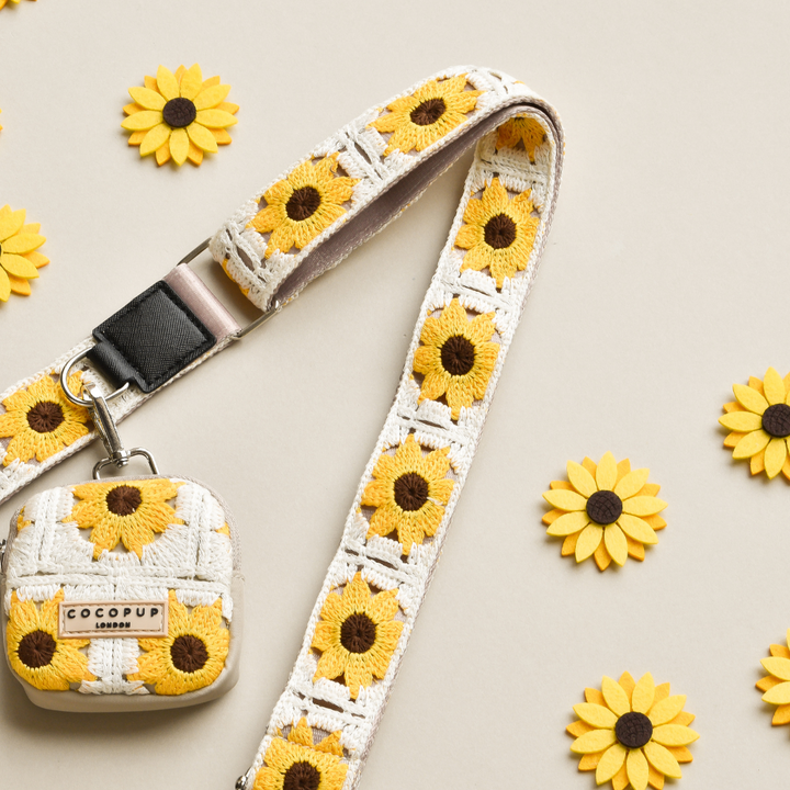 A cream-colored, crochet-style fabric camera strap with yellow sunflower patterns and a matching camera case is displayed on a light beige background. Several small yellow sunflower decorations are scattered around the strap and case, reminiscent of Cocopup London's Sunflower Patch Bag Strap.