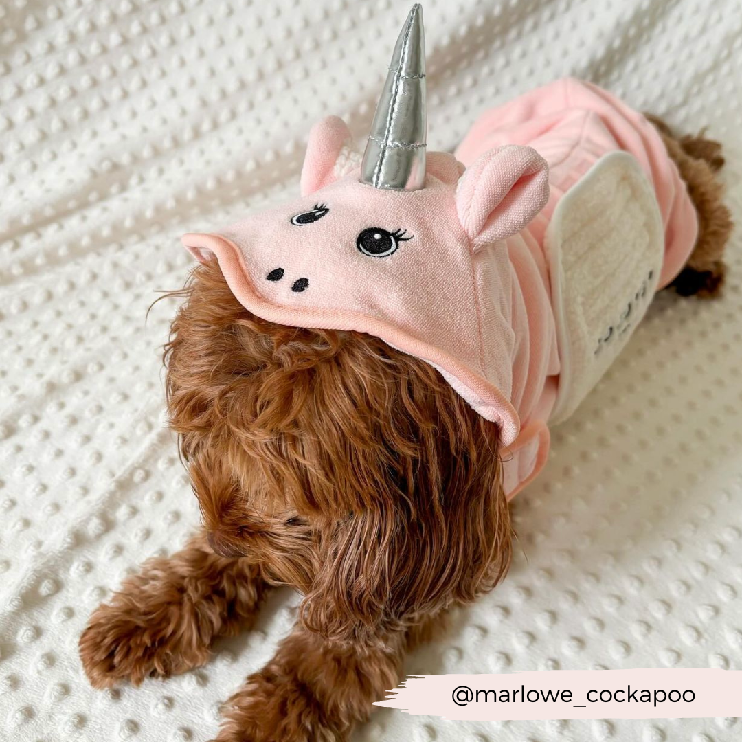 A fluffy brown dog, likely a Cockapoo, is lying down on a textured white blanket. The dog is dressed in the Cocopup London PRE-ORDER Pupicorn Drying Robe, which features a pink unicorn-inspired hood with a silver horn, ears, and an adorable face. The Instagram handle "@marlowe_cockapoo" is visible in the bottom right corner.