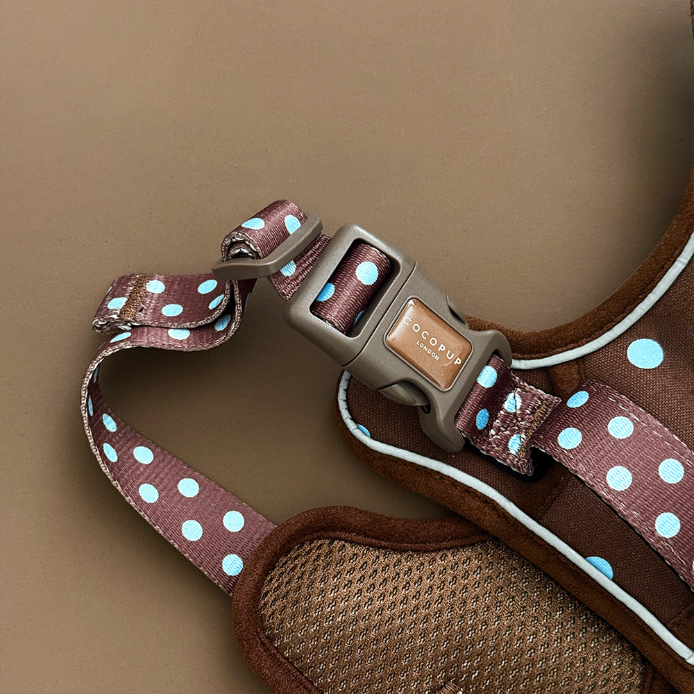 A close up of the buckle on a brown dog harness with light blue polka dots, laid flat on a solid brown background. 