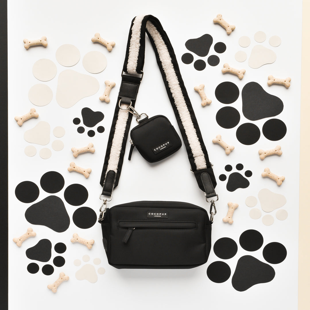 A black crossbody bag featuring the "Bag Strap - Black Aviator" from Cocopup London and a matching mini pouch is displayed on a white background. The arrangement is surrounded by paper cutouts of circles, paw prints, and dog bones, with several small dog biscuits scattered around. Both the pouch and bag proudly feature the Cocopup London branding.