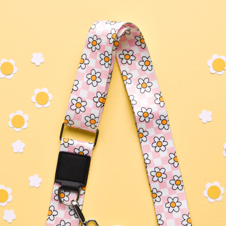 A versatile Bag Strap - Daisy Gingham Rave from Cocopup London featuring a pink and white checkered pattern adorned with yellow and white daisies lies on a light yellow background. Small paper cutout daisies are scattered around the strap.