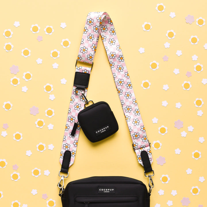 A black bag with a decorative Daisy Gingham Rave strap is displayed on a yellow background with scattered white and pink floral cutouts. A small, black, square pouch featuring the "Cocopup London" brand hangs from this versatile strap, perfect for transforming your bag into a chic Dog Walking Bag.