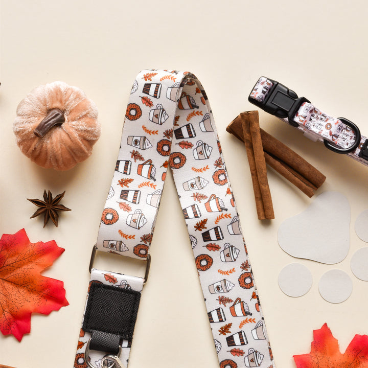 A flat lay of autumn-themed items, including a decorative pumpkin, two cinnamon sticks, star anise, red maple leaves, and the Cocopup London 'Bag Strap - Autumn Puppuccino' featuring patterns of coffee cups and pumpkins on a cream background. Perfect for pairing with your favorite dog walking bag as you sip an autumn puppuccino.