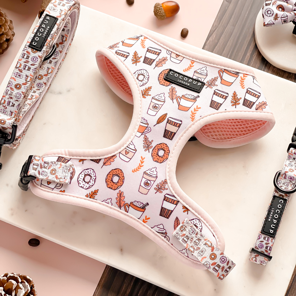 Displayed on a marble surface is the pink Autumn Puppuccino Adjustable Neck Harness from Cocopup London, adorned with autumn-themed patterns such as leaves, donuts, and coffee cups. Surrounding the main harness are partially visible matching accessories from the same bundle, including a collar and lead featuring similar designs.