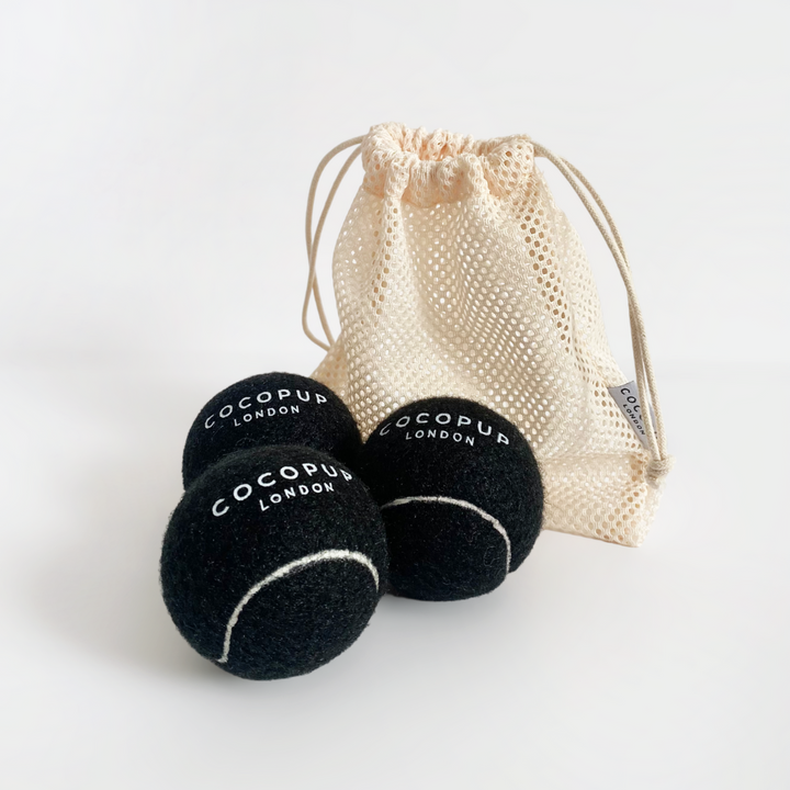 A beige mesh drawstring bag with a small white tag beside three midnight black tennis balls. The tennis balls, perfect as a durable dog toy, have the text "Cocopup London" printed on them in white. The background is plain white.