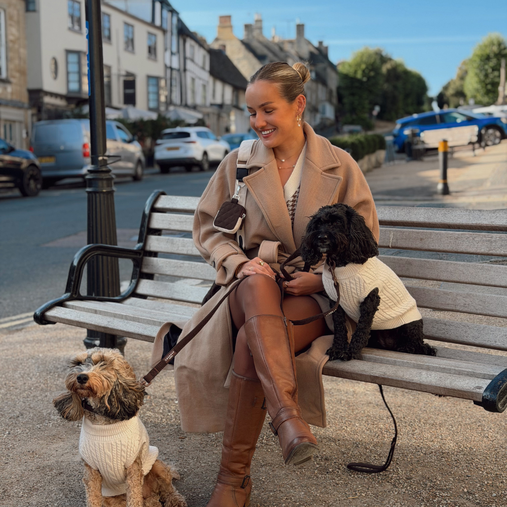 A woman sits on a wooden park bench outdoors with two dogs wearing cream knit sweaters. She is dressed in a camel coat, brown knee-high boots, and a white top, with a black bag slung over her shoulder.