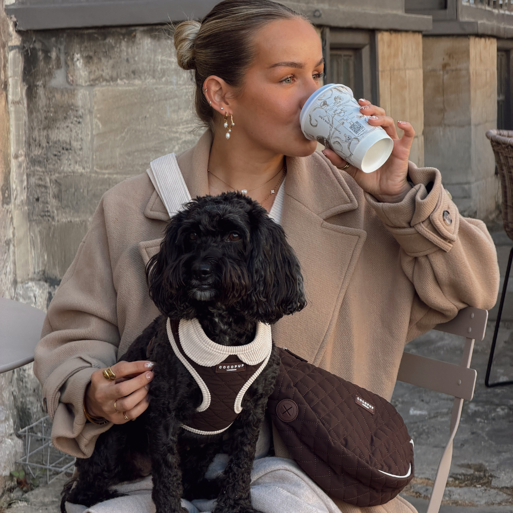A woman in a beige coat sits outdoors holding a black dog wearing a brown and cream dog harness. She has a brown quilted bag carrier bag on her lap and is drinking from a white patterned paper cup.