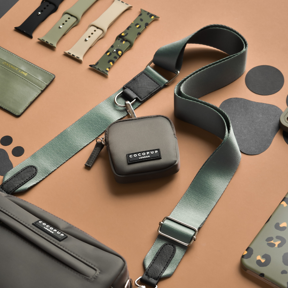 A neatly arranged layout displays green and camouflage-patterned smartwatch bands, a small grey pouch with the Cocopup London "Bag Strap - Khaki," a larger matching Dog Walking Bag, and other accessories on a brown background with black paw print designs.