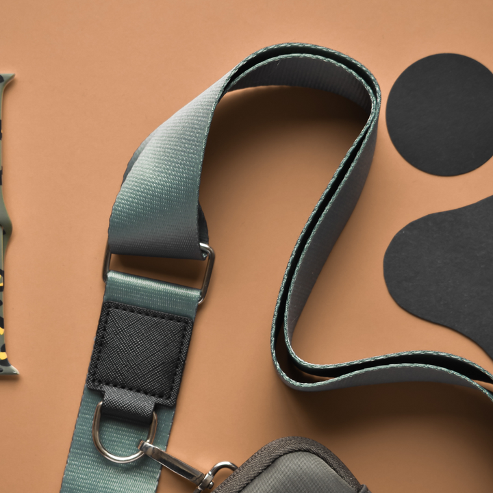 A close-up showcases a green and gray adjustable dog leash featuring a sturdy metal clasp and black fabric accents, all set against an orange-brown background. Also in the scene are a black paw-shaped cutout and part of a matching Cocopup London Bag Strap in khaki.