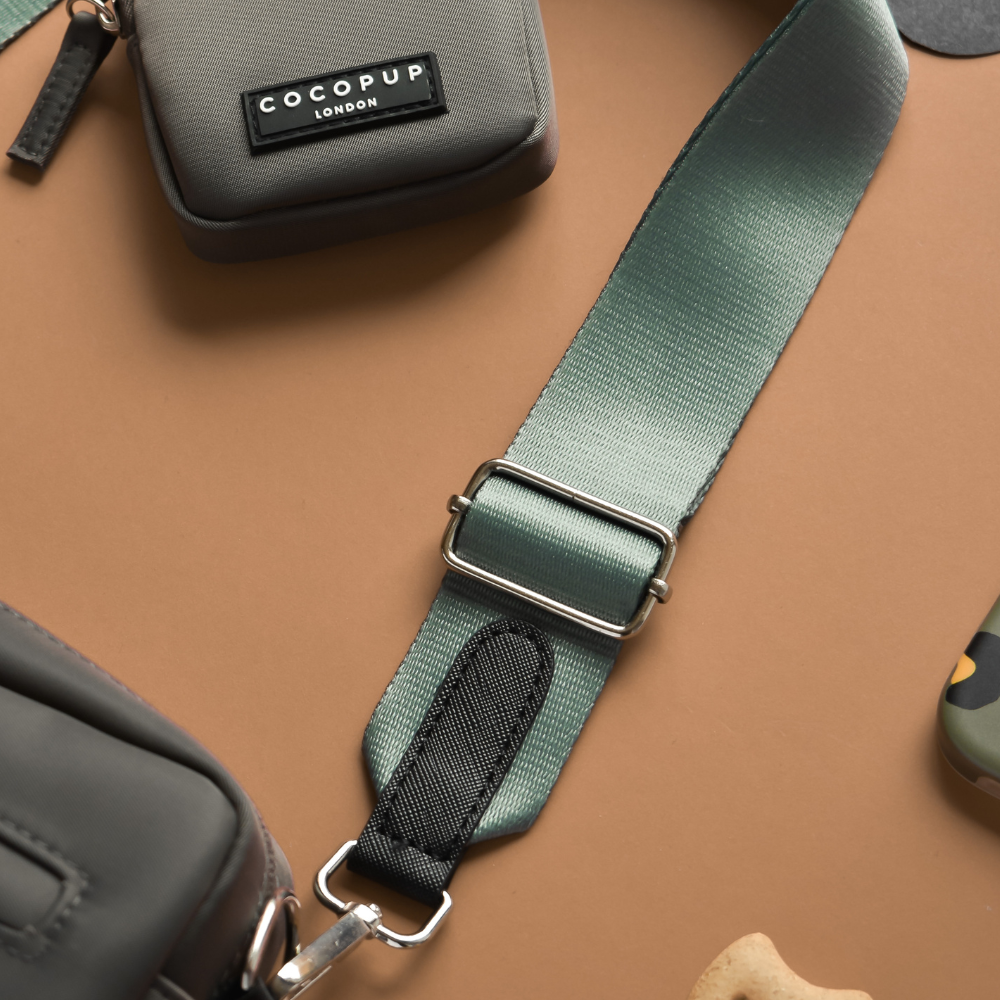 A close-up of stylish accessories on a tan surface highlights a grey dog walking bag with a black "Cocopup London" label, featuring the Khaki adjustable polyester strap with a metal buckle and, partially visible, a camouflage-patterned item that could be a phone case.