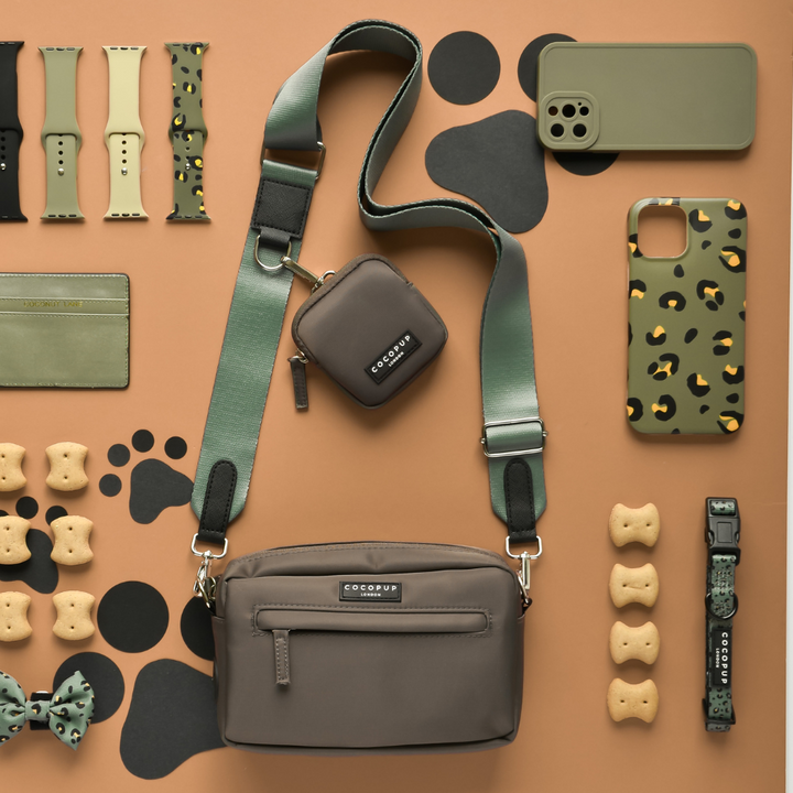 A neatly arranged display of pet accessories, including a crossover dog walking bag, Cocopup London's pouch with a Bag Strap - Khaki, dog treats, a leash, a bow tie, and patterned phone cases. The items are in shades of green and brown polyester with a paw print design on the background.