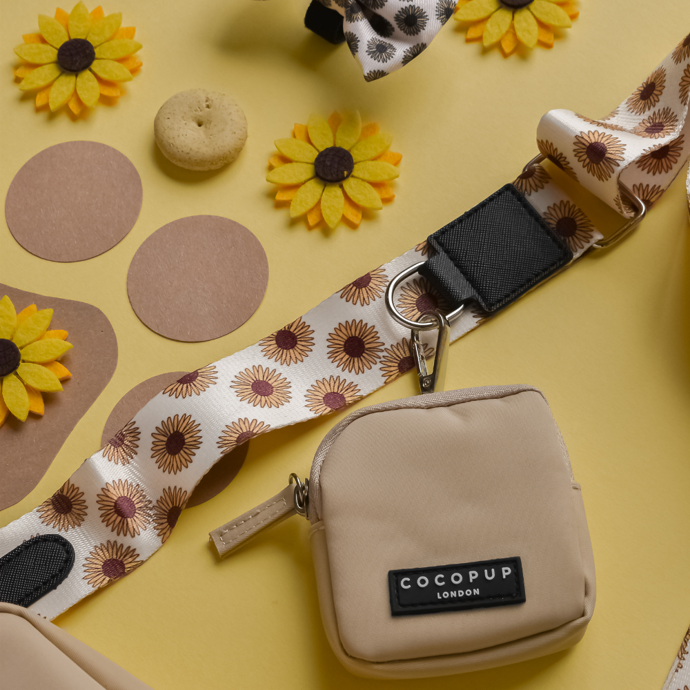 A beige crossbody dog walking bag branded with "Cocopup London" lies on a yellow surface. The bag is attached to a "Bag Strap - Sunflowers" and is surrounded by felt sunflower decorations, beige circles, and a small beige cookie.