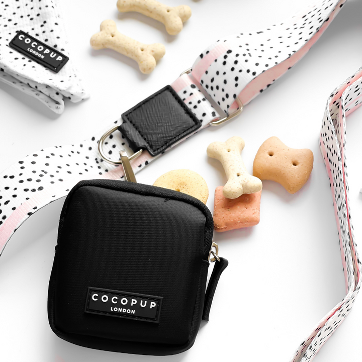 A Black Cocopup London pouch from the Pink Dalmatian Collection is placed beside a white and black spotted Bag Strap - Pink Dalmatian. There are several bone-shaped dog treats scattered around. The background is white and clean, highlighting the dog accessories and treats.