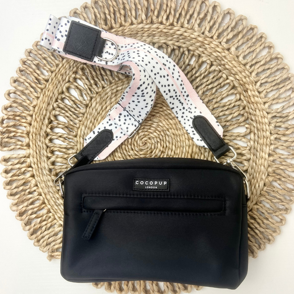 A black Cocopup London Dog Walking Bag with a front zipper pocket is lying on a round, woven straw mat. The bag is attached to a Bag Strap - Pink Dalmatian featuring black leather accents.