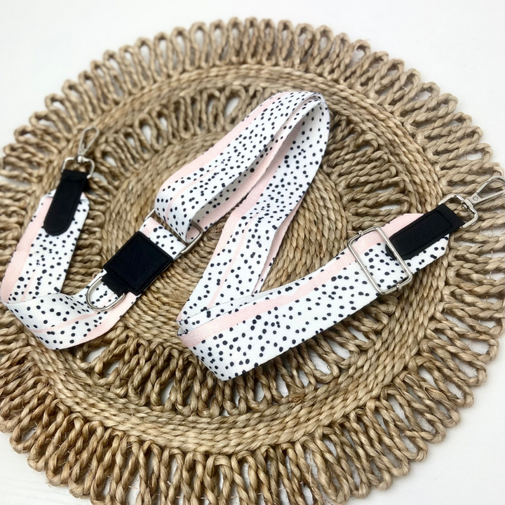 A Bag Strap - Pink Dalmatian from Cocopup London, featuring black polka dots with pink edges, is laid out in a U-shape on a round, woven straw placemat. The white strap has adjustable silver buckles and black leather connectors at the ends.