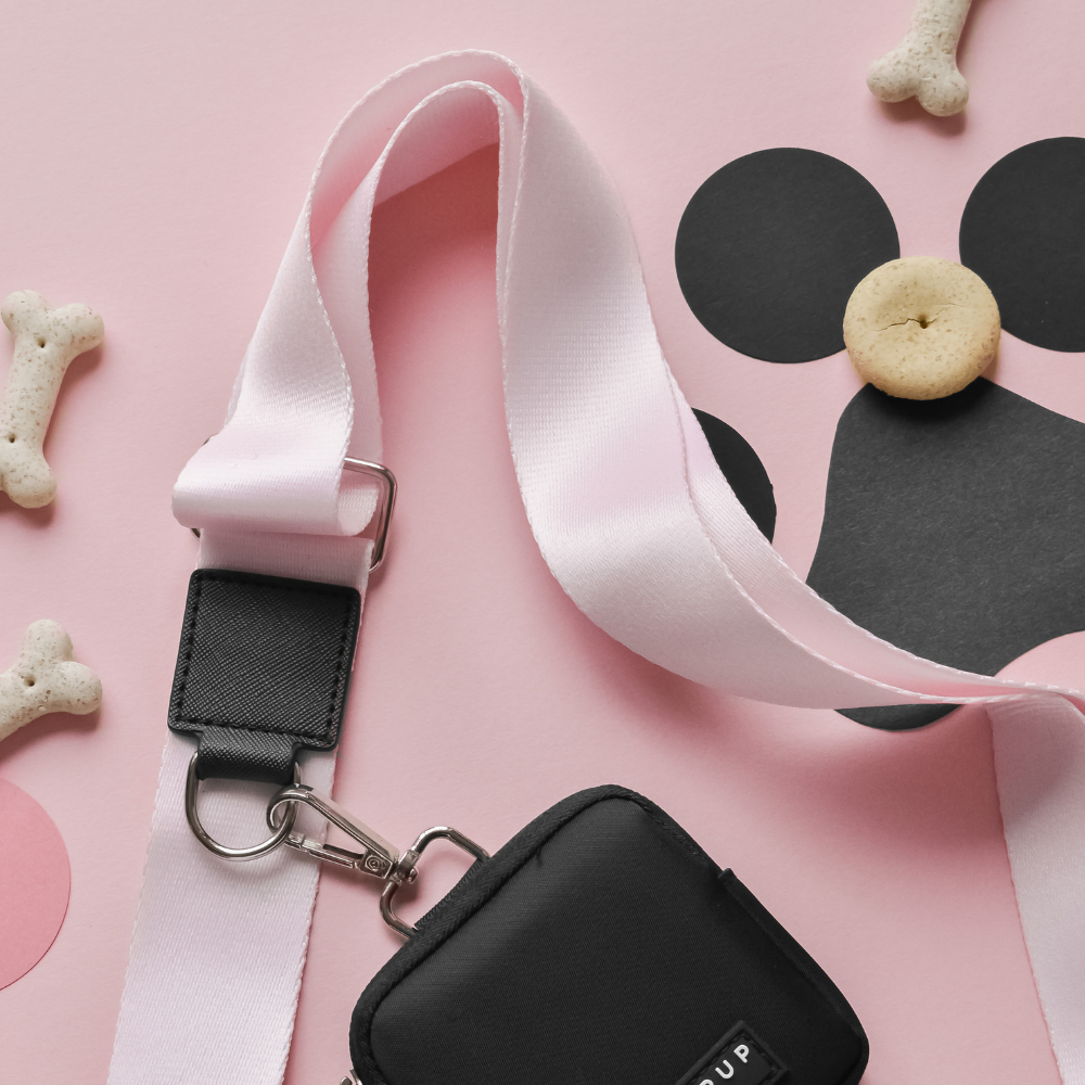 A light pink Bag Strap - Plain Baby Pink from Cocopup London, adorned with a Cocopup Dog Walking Bag, is set against a pink background. Surrounding the strap are black and pink cut-out shapes and dog bone-shaped biscuits, one creatively arranged to resemble a dog face with cut-out pieces—ideal for carrying your everyday essentials.